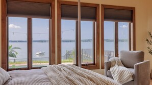 Une chambre avec une grande fenêtre donnant sur un lac, un lit recouvert d’une couverture blanche tricotée et un fauteuil gris.