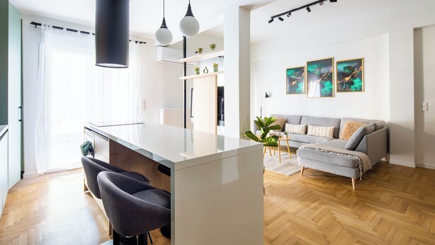 A modern kitchen with a central island, a dining area with a grey sofa, and a living room with a grey sofa and a plant. - Mystique 2-Bedroom Apartment in Athens (Athens)