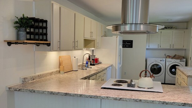 Una cocina moderna con encimera de granito, campana extractora de acero inoxidable y electrodomésticos integrados. Hay dos taburetes de madera y una pequeña isla con un hervidor.