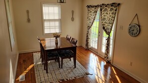 Un comedor con una mesa de madera, sillas, un jarrón con flores, un tapete estampado y una ventana con persianas.