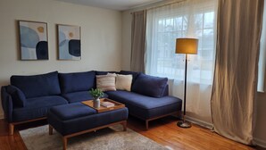 Un salón con un sofá seccional azul oscuro, una mesita de centro de madera con un jarrón y un libro, una alfombra estampada y una lámpara de pie.