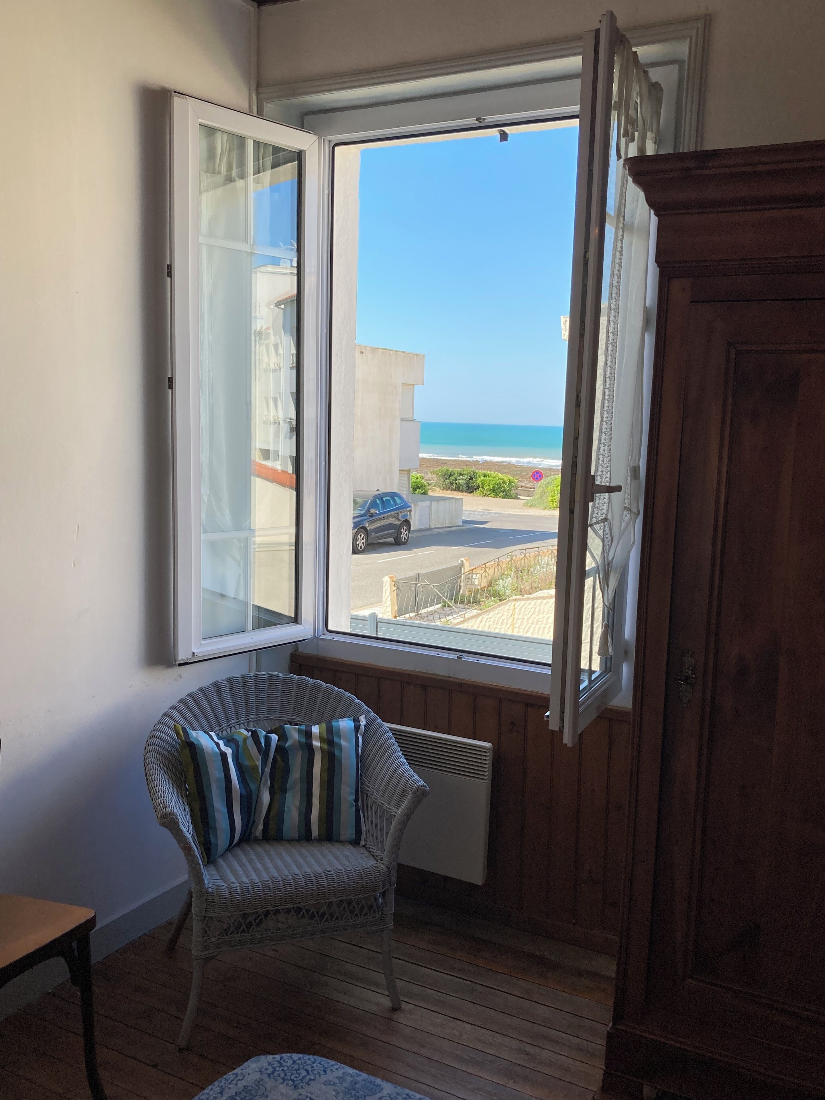 Une pièce avec un fauteuil en osier, une armoire en bois et une fenêtre donnant sur la plage et une voiture garée.