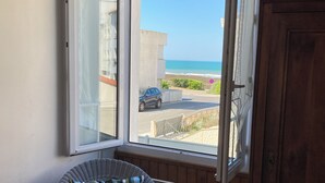A room with a wicker chair, a wooden cabinet, and a window with a view of the beach and a parked car.