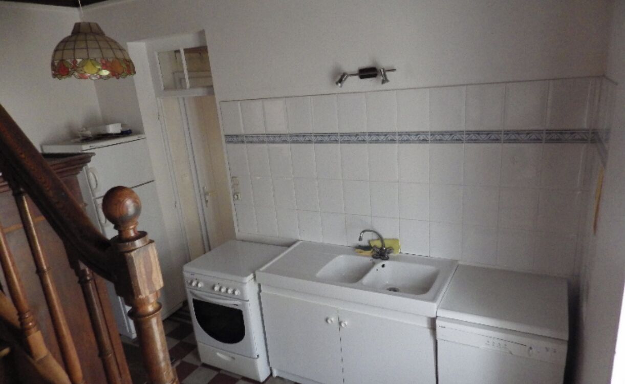 Ett kompakt kök med vita vitvaror, en trätrappa och en kakelbeklädd vägg bakom diskbänken.