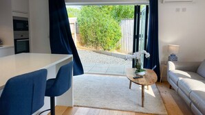 Et moderne køkken med hvid bordplade og indbygget ovn, et spiseområde med blå stole, et rundt træbord med en vase med hvide blomster og en sofa med en lampe.