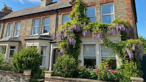 A two-story house with a brick facade, a pitched roof, and a front garden with flowering plants and hanging purple flowers. - Spacious Five Bed Home in the Heart of Cambridge (Cambridge)