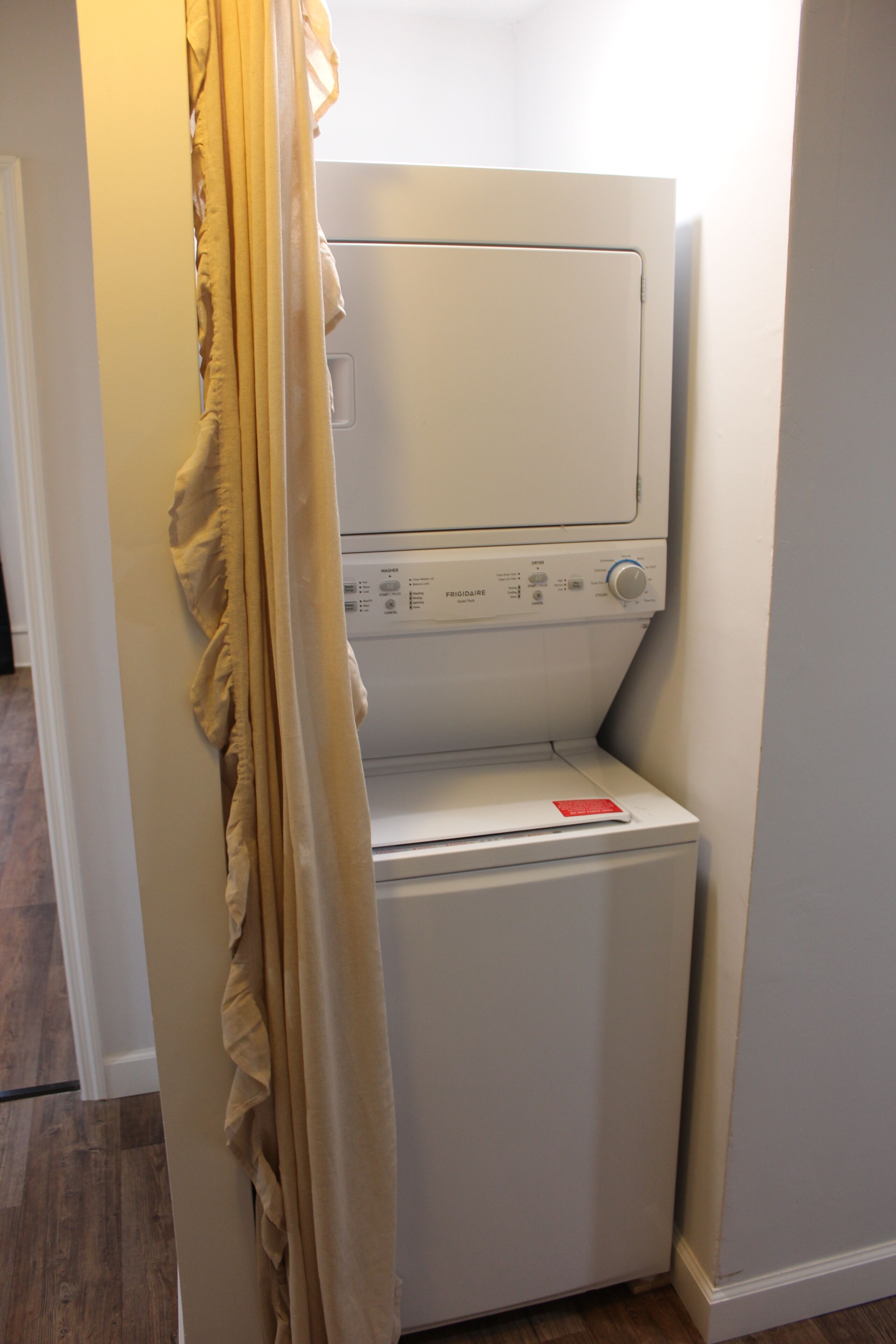A compact, white laundry machine in a corner of a room with wooden flooring and a plain wall.