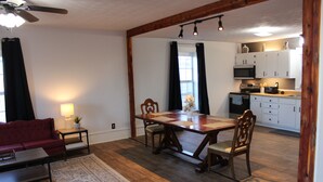 A dining area with a wooden table and chairs, a red sofa, and a small rug. A kitchen with white cabinets and appliances is visible in the background. - Charming historic home steps from downtown LaGrange (LaGrange)