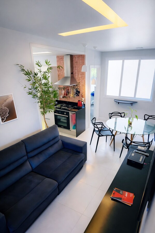 Una sala de estar moderna con un sofá azul oscuro, un comedor con mesa y sillas de vidrio, una cocina con baldosas rojas y un ventanal que proporciona luz natural.