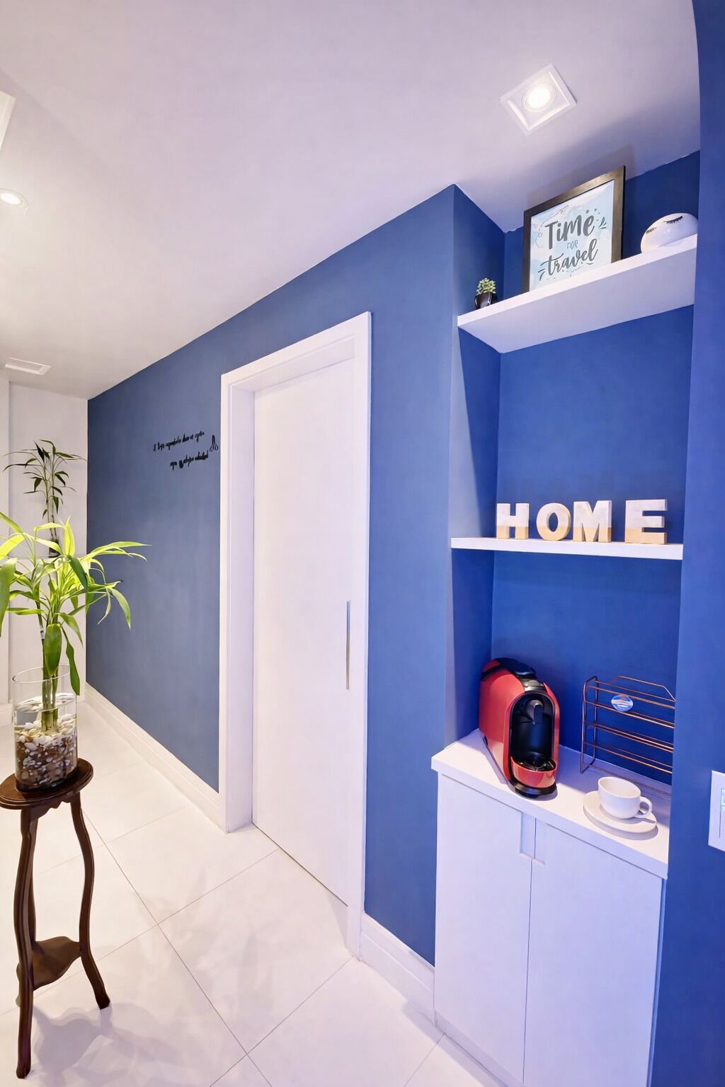 Un pasillo con una pared azul decorativa, una puerta blanca, un estante con la palabra “HOME” y un objeto rojo, y una planta sobre una mesita.