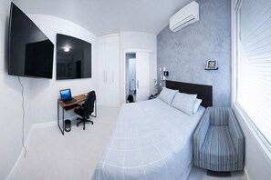Un dormitorio moderno con cama, escritorio con laptop, televisor y aire acondicionado.