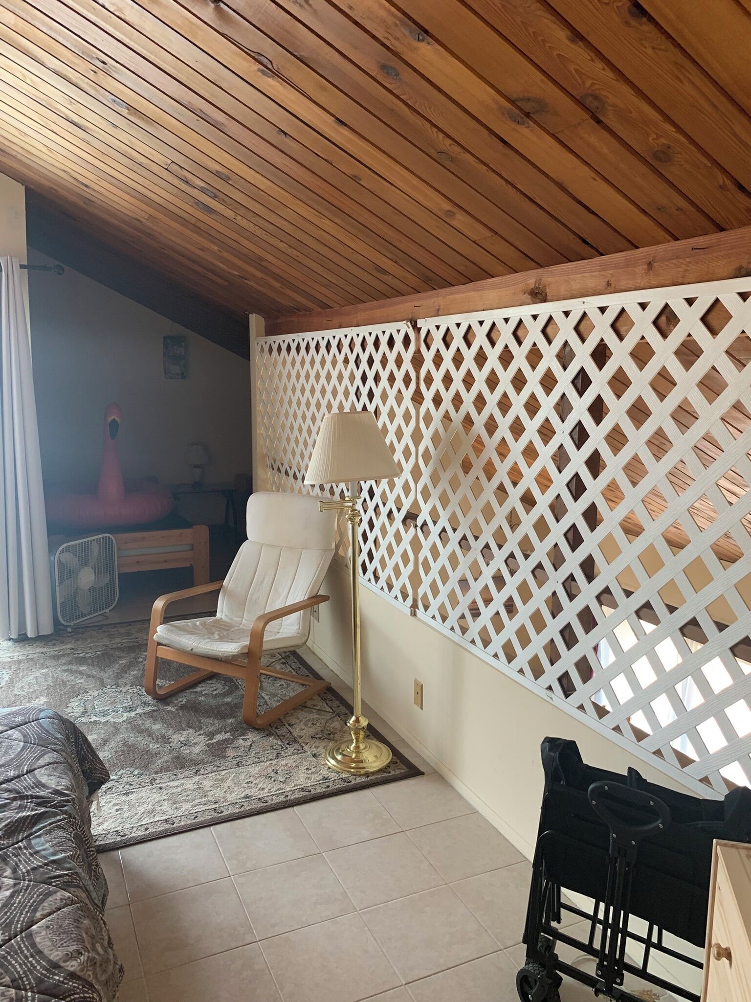 Et værelse med træloft, en hvid krydsramsestruktur, en hvid stol, en gulvlampe, en sort klapvogn og en stribet sofa.