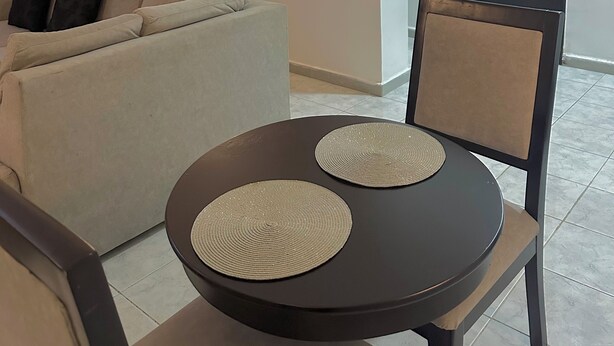 A modern living room with a sofa, a round table with two placemats, a wall-mounted light, and a door. - Charming 2-bedroom apartment with AC, WiFi in the Heart of Wuse II, Abuja. (Abuja)