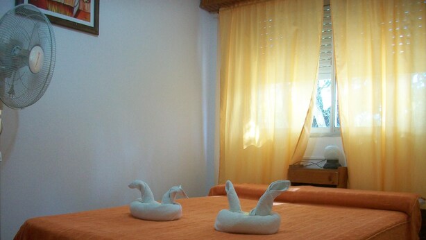 A hotel room with two beds, an orange bedspread, a fan, a window with yellow curtains, and a framed picture on the wall. - Hotel Gli Amici (Mar de Ajo)