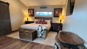 A bedroom with a bed, wooden headboard, bedside tables with lamps, a dresser, a vanity with a mirror, and a window with a view. - Rest your Kicks off of Route 66 at Rain Valley Inn (Flagstaff)