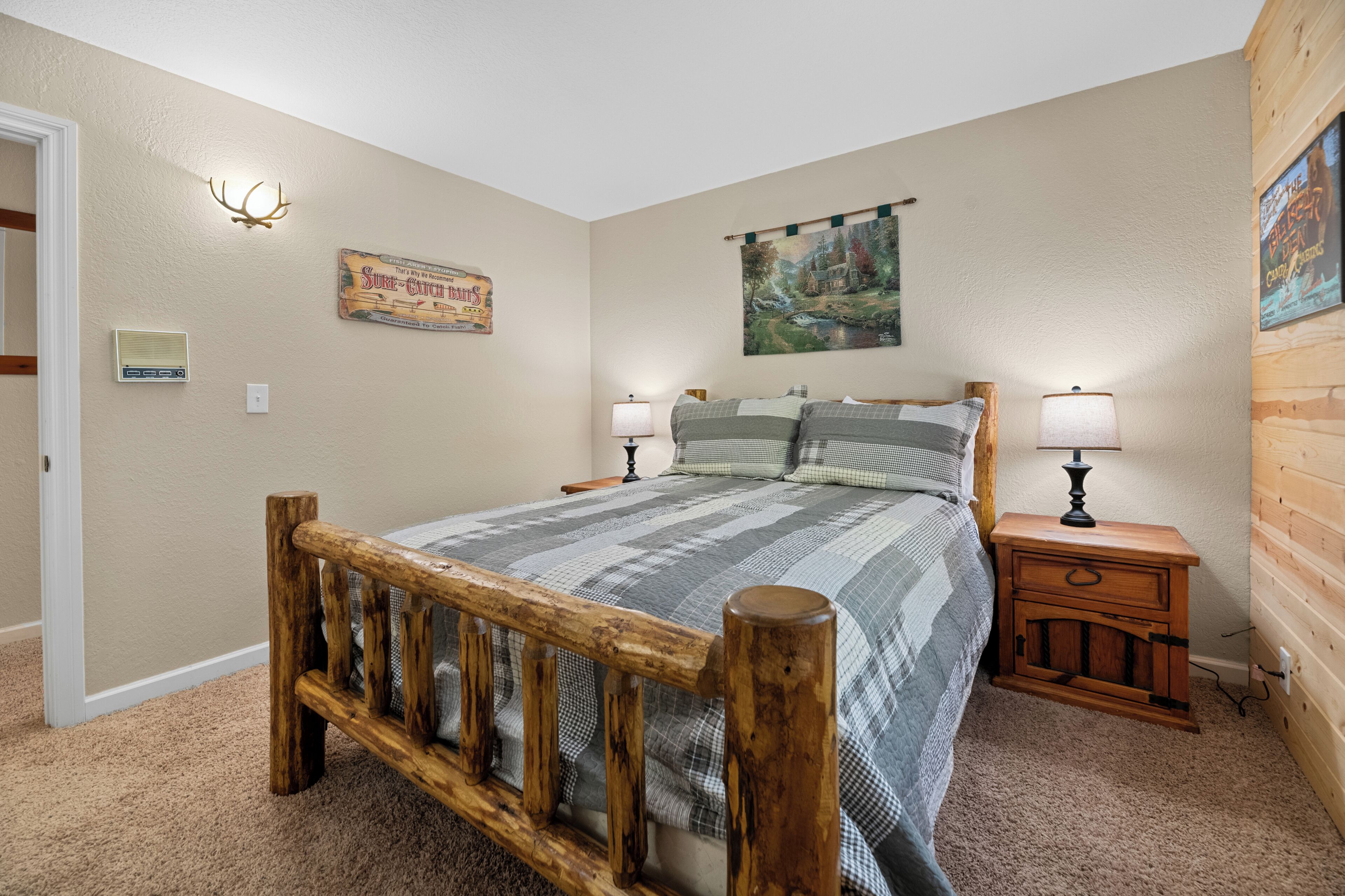 A bedroom with a wooden bed frame, a checkered bedspread, two bedside tables with lamps, a wall-mounted heater, a poster, and a painting.