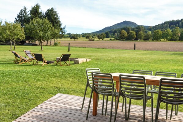 Una terraza de madera con una mesa y sillas, rodeada por un césped con árboles y una colina al fondo.