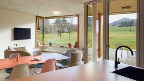 Una cocina moderna con un ventanal grande, un comedor con mesa y sillas, y vistas a un campo verde y montañas.