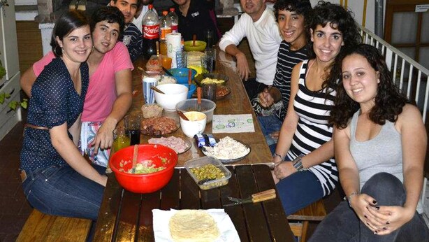 A group of people seated around a wooden table with various food items and drinks, in a room with a brick wall and a window. - La Casa del Girasol Residencia Universitaria (Buenos Aires)