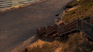 Una playa con una escalera de madera que desciende a la arena, un cielo despejado y el sol poniéndose sobre el agua.