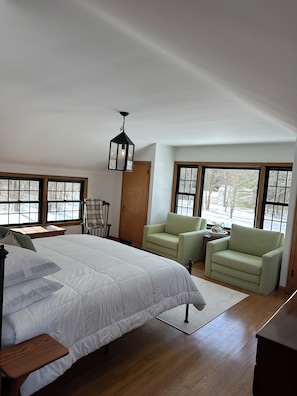 Une chambre à coucher avec un grand lit, deux fauteuils verts, une porte en bois et de grandes fenêtres.