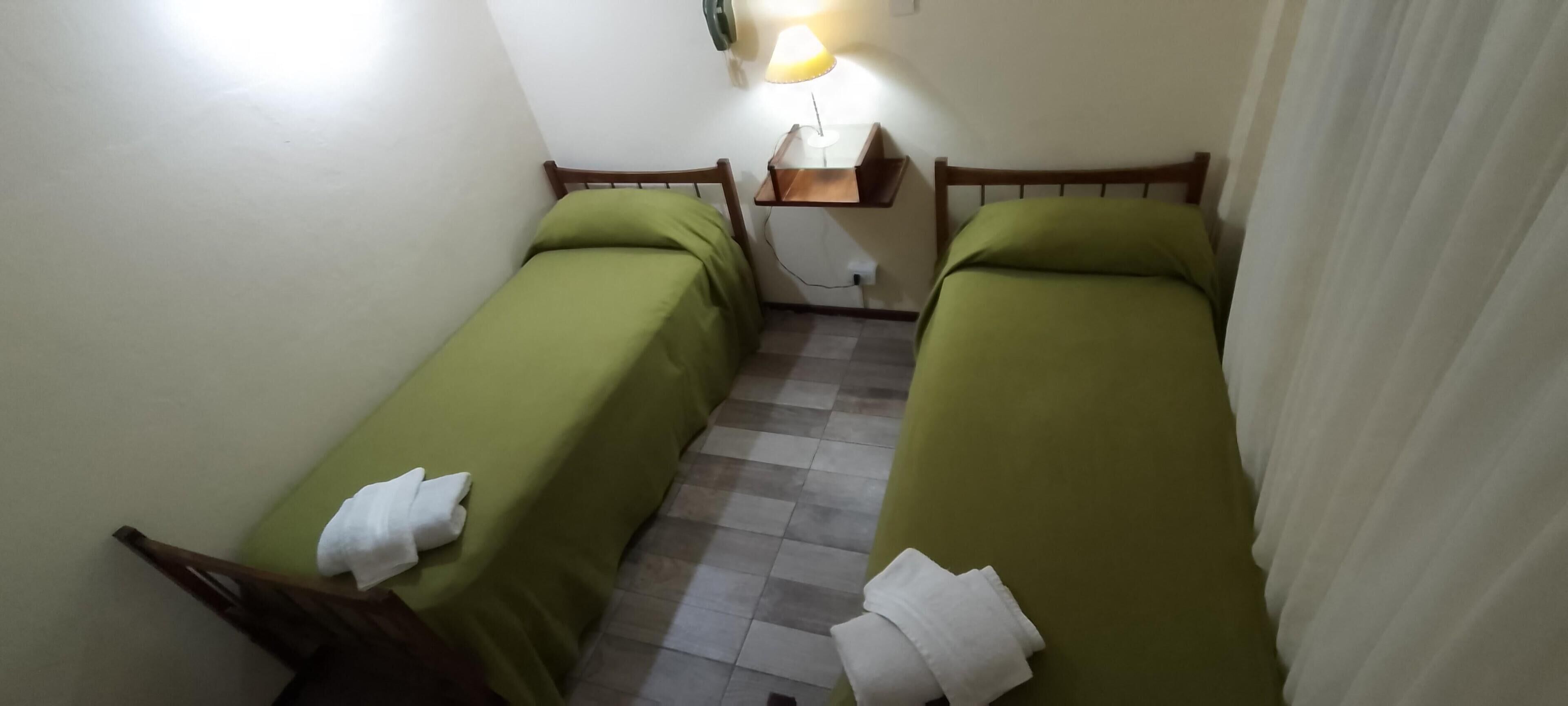 Une petite pièce faiblement éclairée, avec deux lits simples, une table de chevet, une lampe et une chaise.