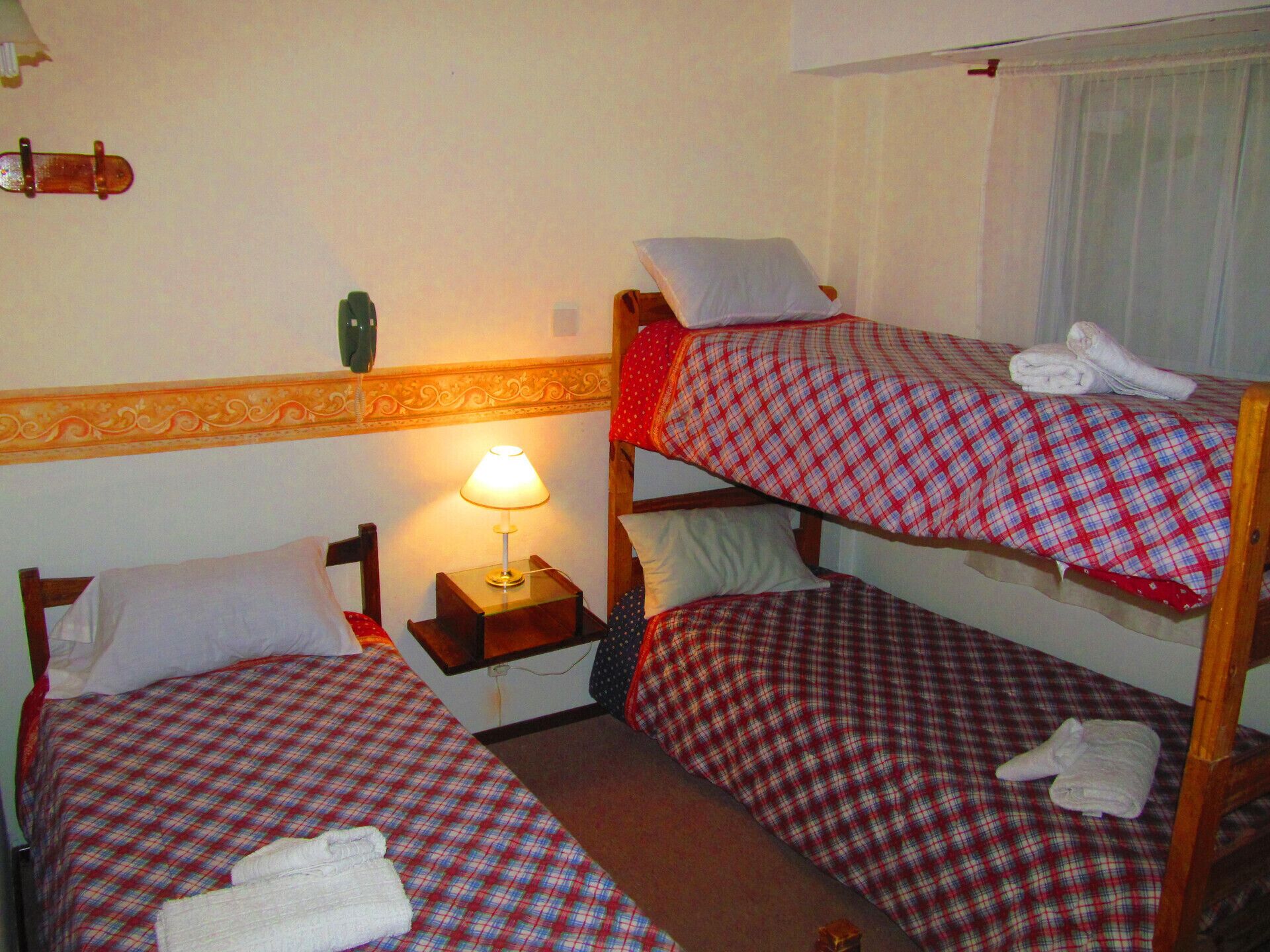 A hotel room with two single beds, each with a plaid bedspread and pillows, a nightstand with a lamp, and a small table with a vase.
