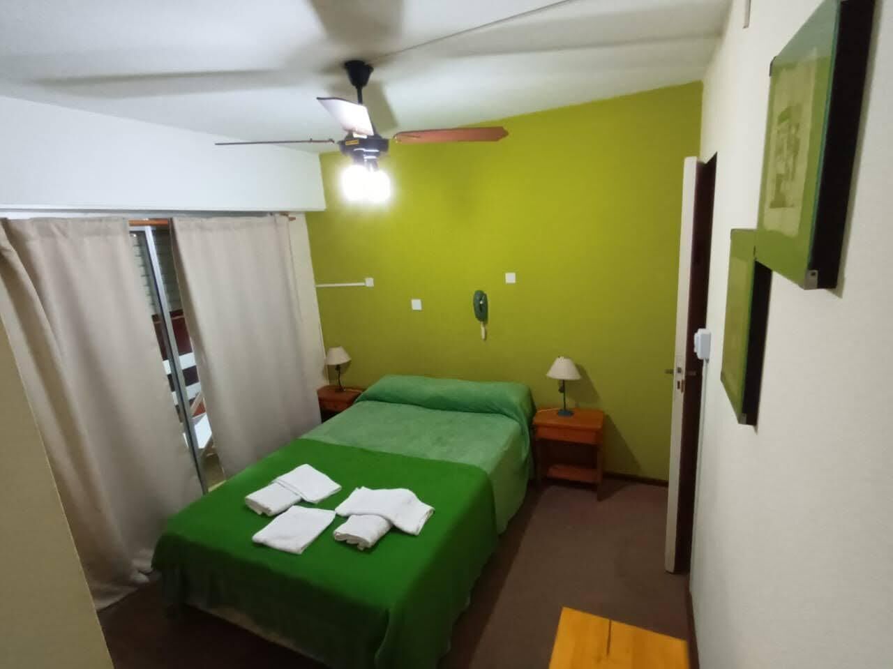 A small, single-bed room with a green bedspread, a ceiling fan, two bedside lamps, and a door with a mirror.
