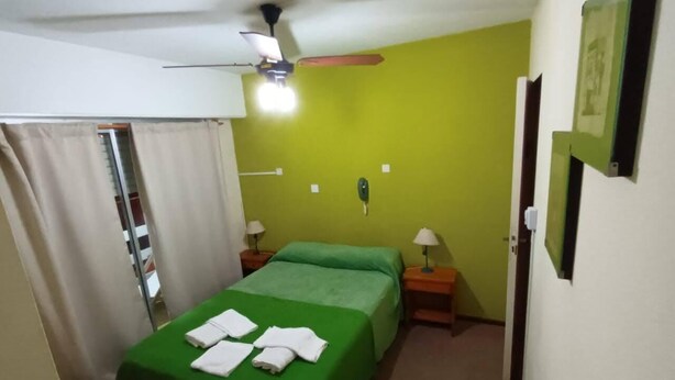 Ein kleines Zimmer mit einem Einzelbett, einer grünen Tagesdecke, einem Deckenventilator, zwei Nachttischlampen und einer Tür mit Spiegel.