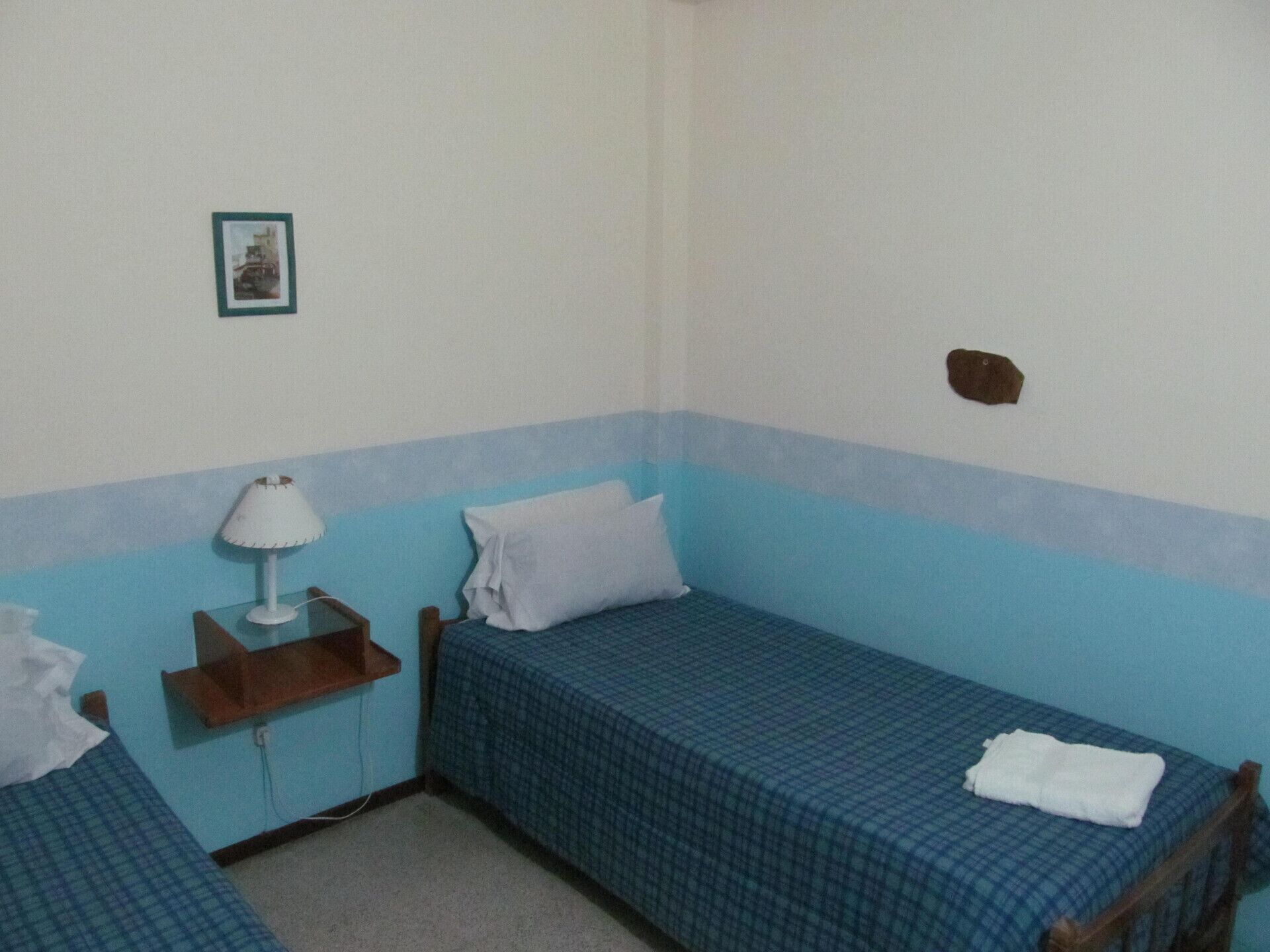 A small, single-room accommodation with two beds, a bedside table with a lamp, and a wall-mounted shelf.
