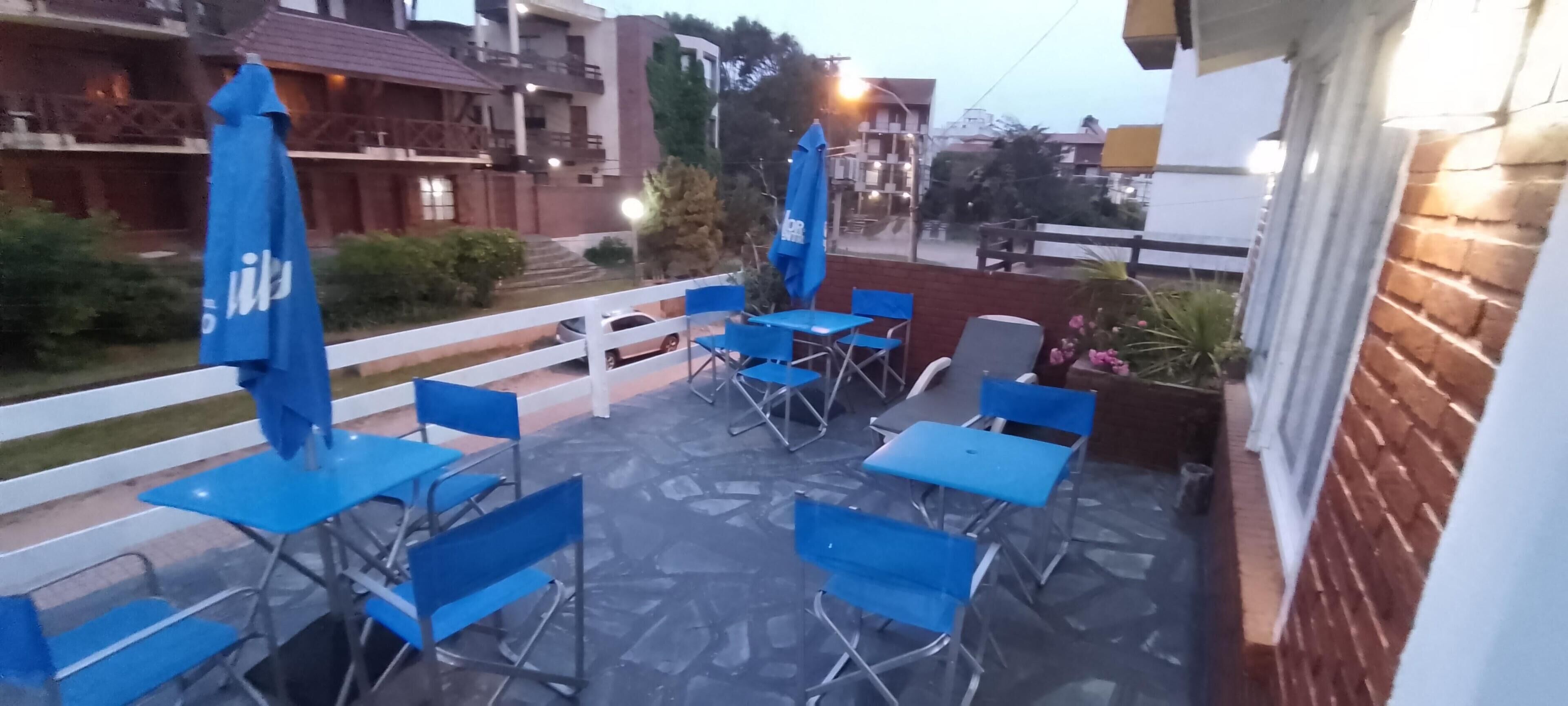 Een terras buiten met blauwe klapstoelen en tafels, een wit hek en een bakstenen muur.