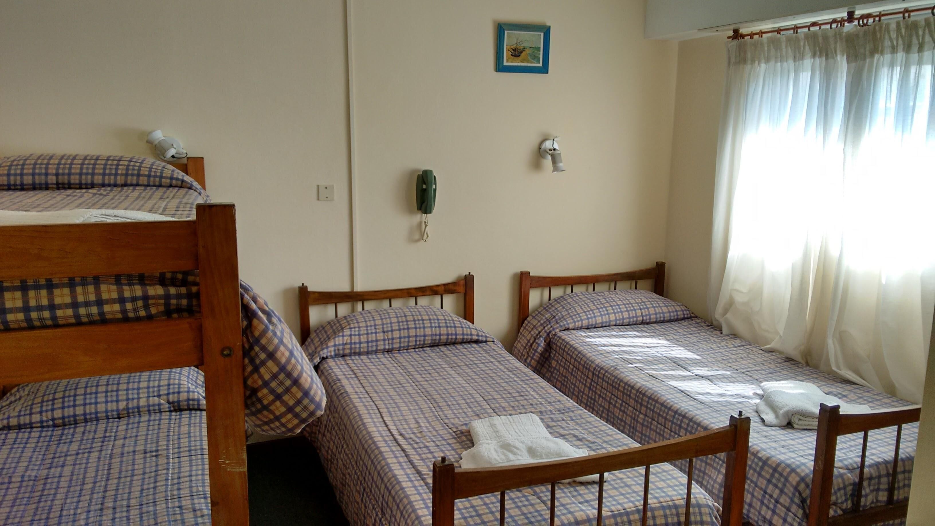 Un dormitorio con dos camas individuales y una litera, todas con ropa de cama escocesa. Hay una pequeña foto en el marco en la pared, un teléfono y una ventana con cortinas traslúcidas.