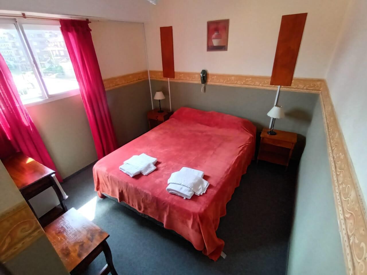 A hotel room with a bed, a desk, a nightstand, a lamp, and a window with curtains.