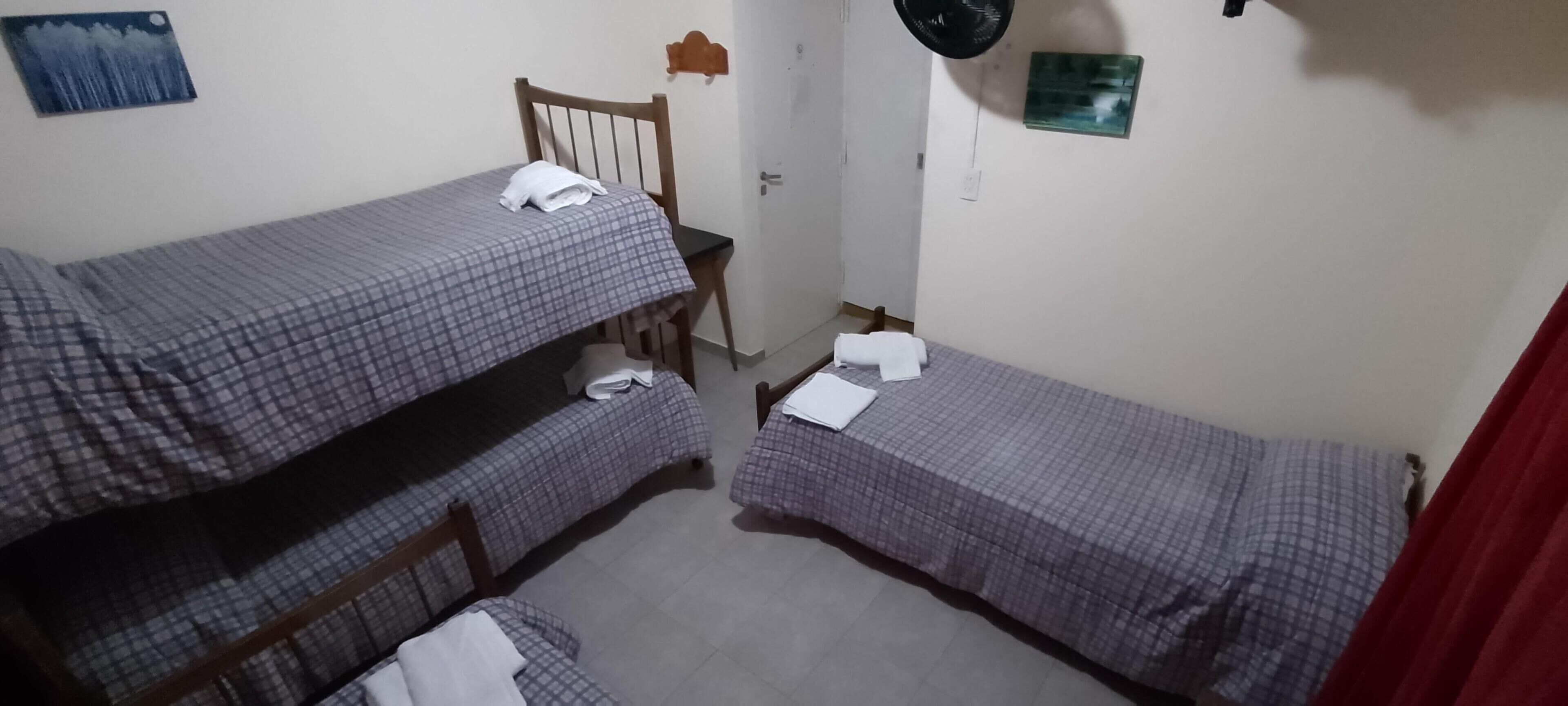 A dormitory room with two bunk beds, a wooden chair, a small table, and a wall-mounted fan.