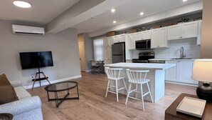 Una sala de estar moderna con sofá, mesa de centro y televisor, comunicada con una cocina que tiene armarios blancos y una isla central.