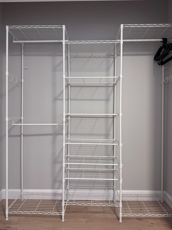 A white metal shelving unit with multiple shelves, likely used for storage in a room with wooden flooring and a plain wall.