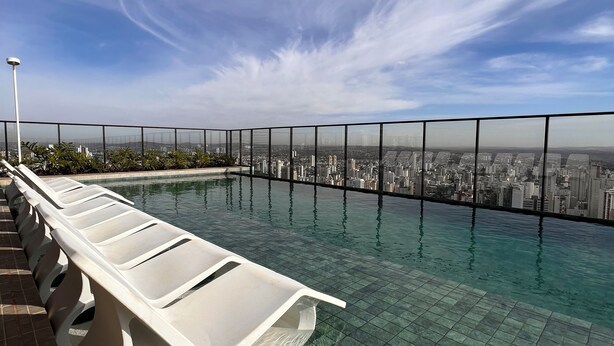 An infinity pool with a city view, surrounded by a glass railing and a tiled deck. - ID URBAN LIFE LUXURY STUDIO FOREST (Setor Oeste Goiânia)