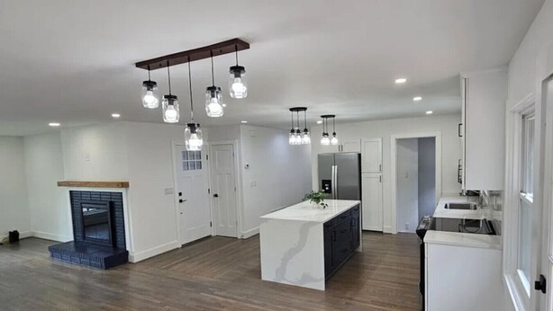 A spacious kitchen with a central island, a built-in oven, and a refrigerator. There is a fireplace with a wooden mantel and a blue accent piece. The kitchen has white cabinets and a wooden floor. - Downtown Oak City Oasis (Raleigh)