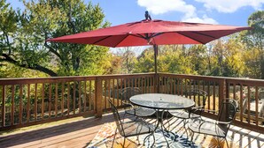 Een houten terras met een ronde tafel en stoelen, een rode parasol en uitzicht op bomen.