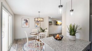 Een moderne keuken met granieten aanrechtbladen, witte kasten en een eethoek met een ronde tafel en stoelen. Er hangen hanglampen boven het keukenwerkblad en een groot raam zorgt voor natuurlijk licht.