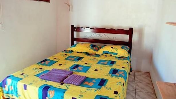 A small, single-bed room with a wooden headboard, a colorful bedspread, and a small window with wooden shutters. - Pousada Casa Castelo Bed e Breakfast (Aracati)