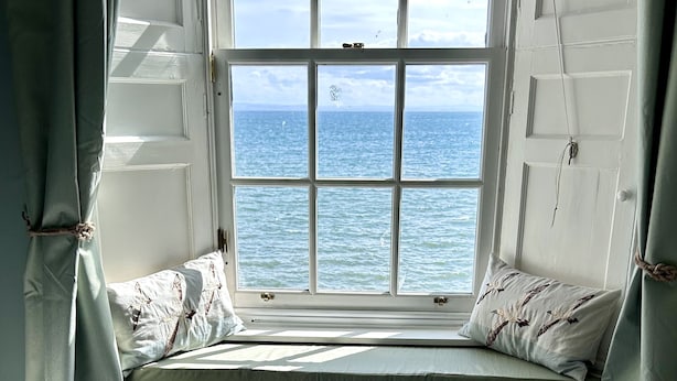 A room with a bed, a window seat with pillows, and a view of the sea.