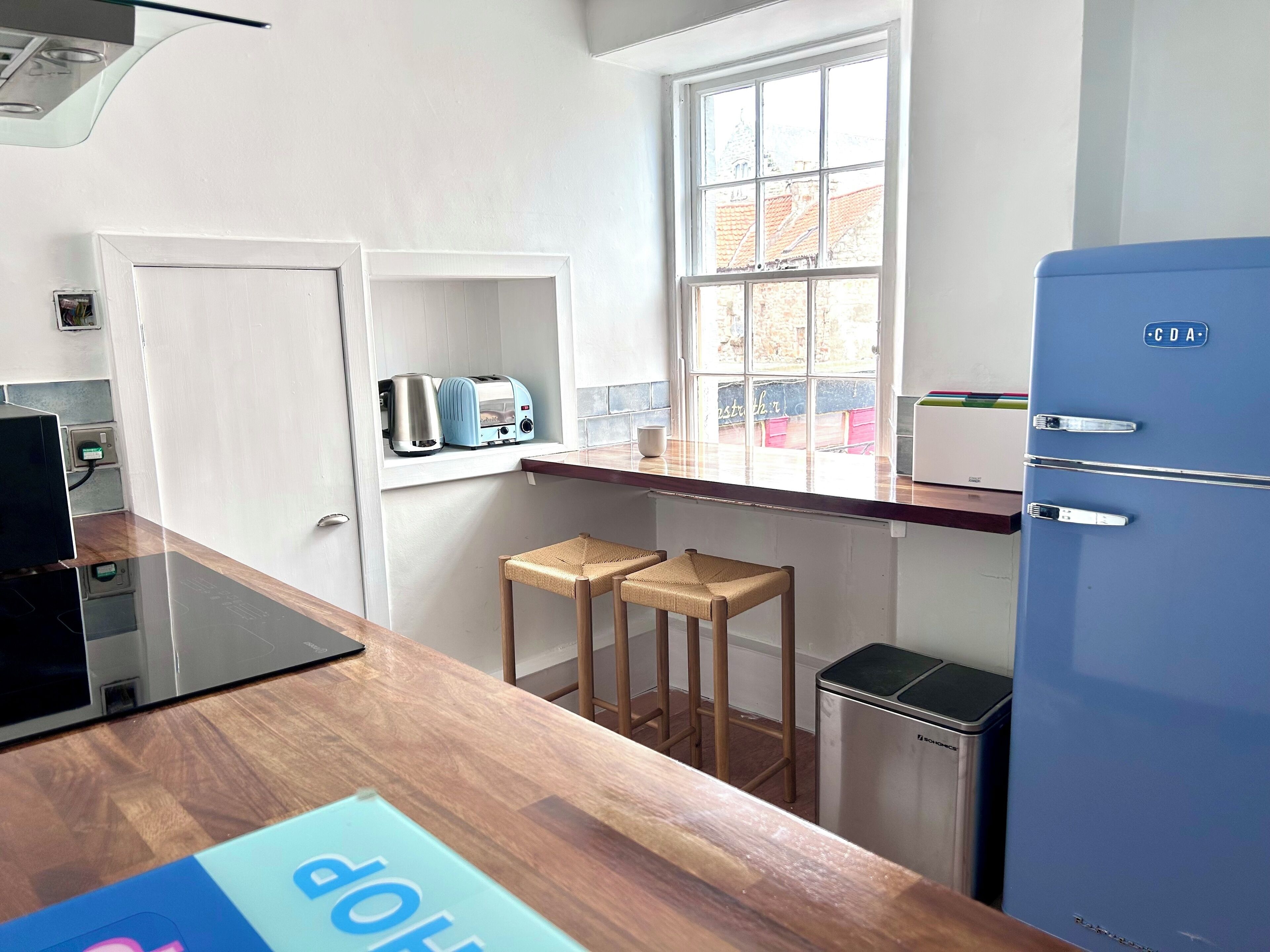 Een kleine keuken met een blauwe koelkast, een raam met uitzicht op gebouwen, een broodrooster en een waterkoker op een plank.