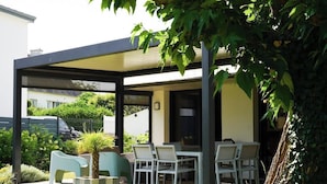 A covered outdoor seating area with a table and chairs, surrounded by greenery and a tree.
