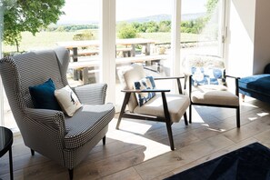 A room with a houndstooth patterned armchair, a beige armchair with a blue pillow, and a blue sofa. There is a wooden floor and a view of a garden through large windows. - Disdow House -  a house that sleeps 11 guests  in 5 bedrooms (Gatehouse of Fleet)