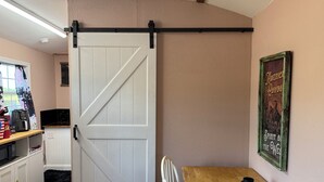 Een kamer met een schuifdeur in landhuisstijl, een houten tafel, een stoel, een keuken met apparatuur en een aan de muur bevestigd fotolijstje.