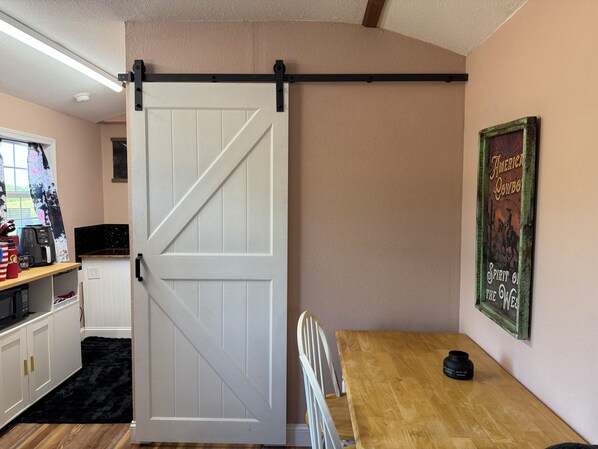 Una habitación con puerta corredera estilo granero, una mesa de madera, una silla, una zona de cocina con electrodomésticos y un cuadro colgado en la pared.