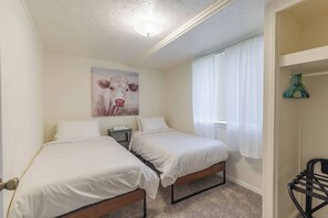 Ein kleines, sauberes Zimmer mit zwei Einzelbetten, einem Nachttisch, einem Bild von einer Kuh und einem Fenster mit durchsichtigen Vorhängen.
