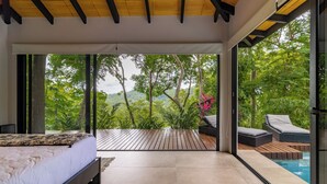 A modern bedroom with a large bed, a wooden deck, and a view of lush greenery and a pool. - SANTA TERESA SURFING VILLA N°2 PRIVATE SWIMMING POOL OCEAN VIEW (Puntarenas Province)