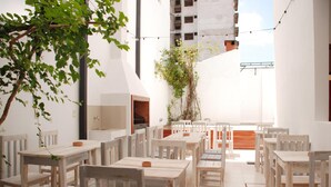 Comedor al aire libre con mesas y sillas de madera, rodeado de paredes blancas y un árbol.